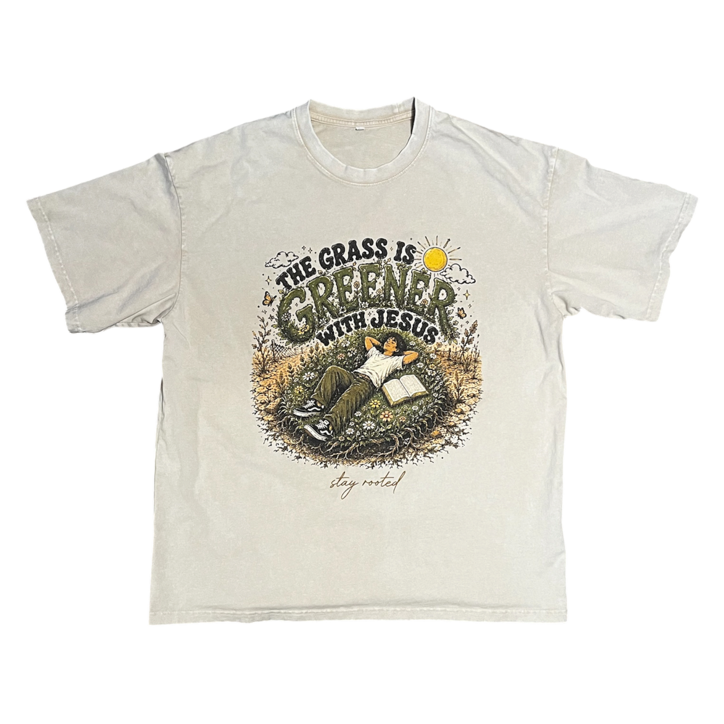 A light beige crewneck t-shirt featuring a graphic print on the front with the text 'THE GRASS IS GREENER WITH JESUS' and an illustration of a person reading in a grassy field.
