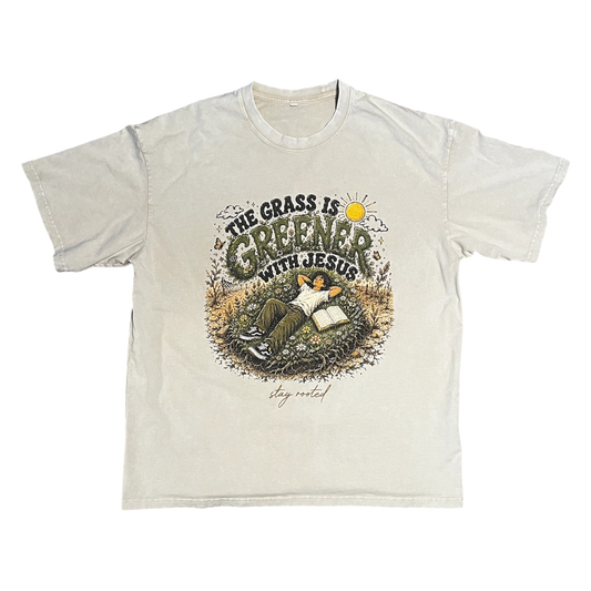A light beige crewneck t-shirt featuring a graphic print on the front with the text 'THE GRASS IS GREENER WITH JESUS' and an illustration of a person reading in a grassy field.